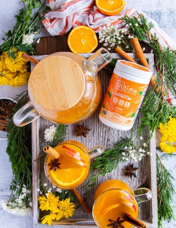 A tray with the large batch of homemade apple cider in a pitcher and two cups served up and garnished with whole spice, oranges and apples, flowers and a tea towel
