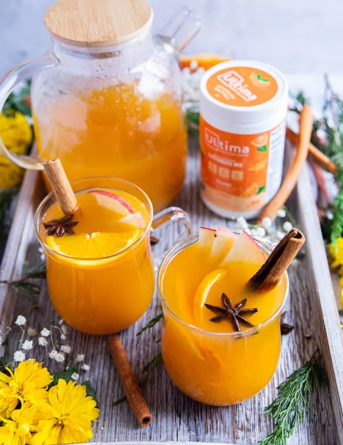 A tray with two cups of homemade apple cider and a large glass jar of apple cider to serve extras, surrounded by flowers, and garnished with cinnamon sticks, orange slices, apple slices