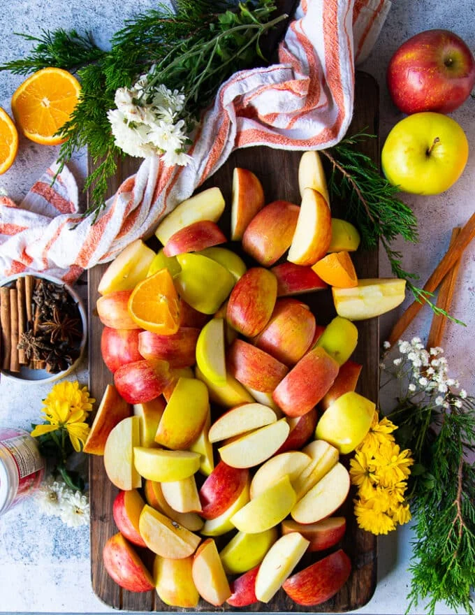 ingredients for apple cider including lots of quartered apples, a plate of spices, oranges cut in quarters, an orange sweetener
