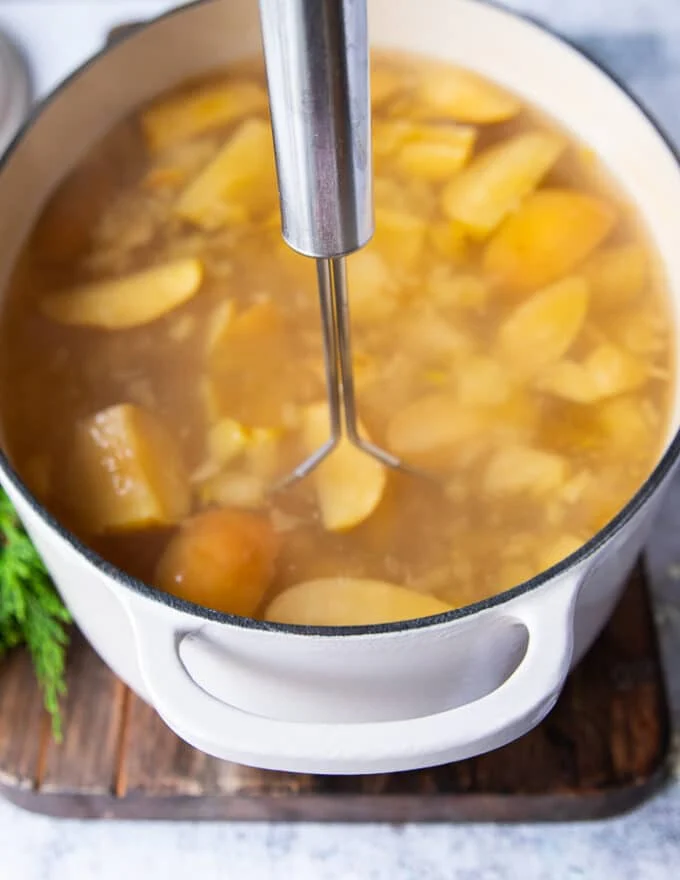 A hand holding a potato masher and mashing down the apples and oranges in the pot to make homemade apple cider