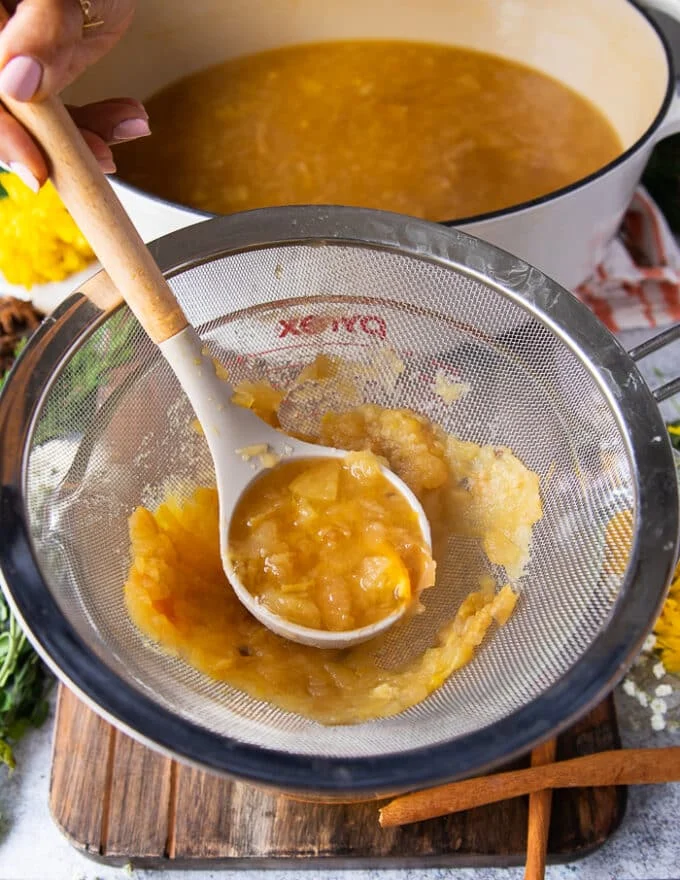 a hand holding a ladle of homemade apple cider and placing it over a fine mesh strainer
