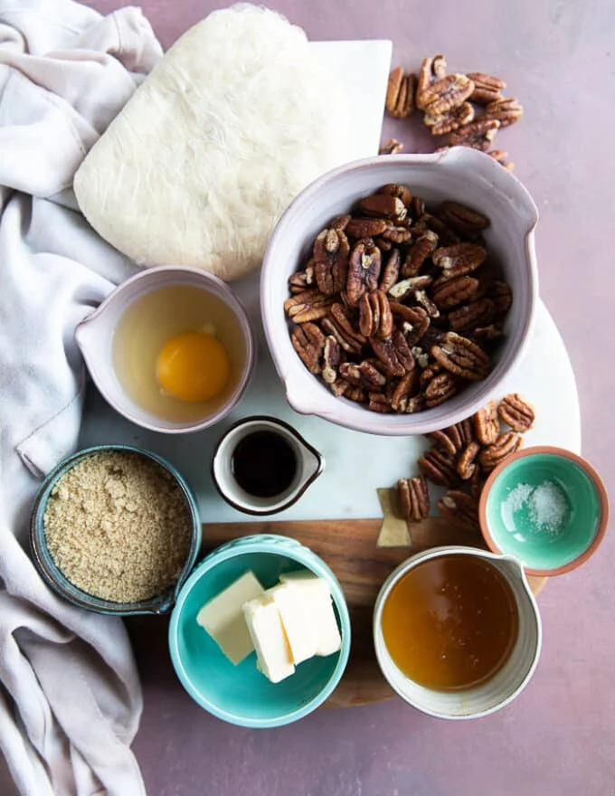 ingredients to make mini pecan pies including a bowl of pecans, a small bowl of honey, brown sugar, one egg, a small bowl of vanilla, another of salt and a cold pie dough wrapped in plastic