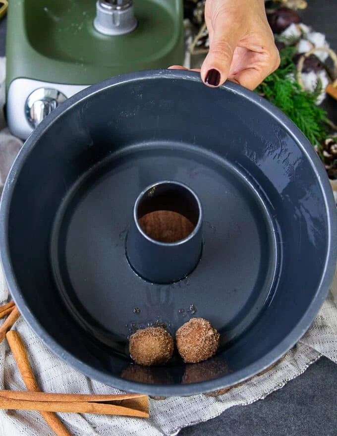 a sprayed bundt pan and a hand arranging the dipped and coated cinnamon roll monkey bread pieces at the bottom of the pan