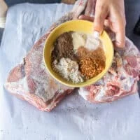 A hand holding a dry spice mix over fresh leg of lamb
