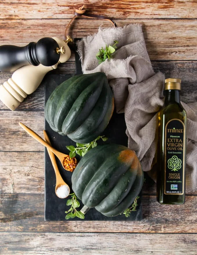 Two washed and cleaned acorn squashes on a black board surrounded by olive oil, salt and pepper shakers