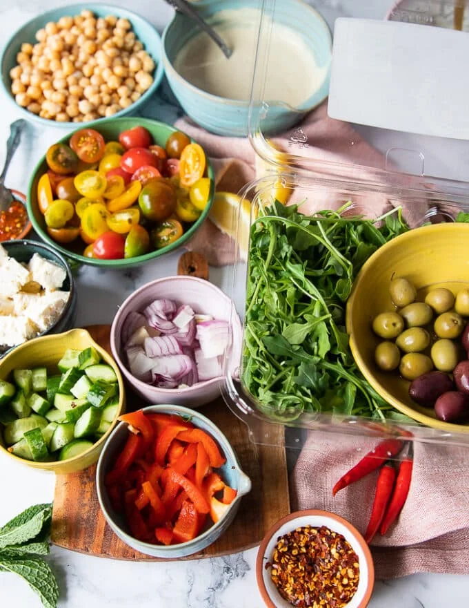 pita bowl topping ingredients: arugula, chickpeas, olives, tomatoes, sweet peppers, chilli peppers, onions, cucumbers, feta, tahini sauce, yogurt sauce, herbs mint and dill
