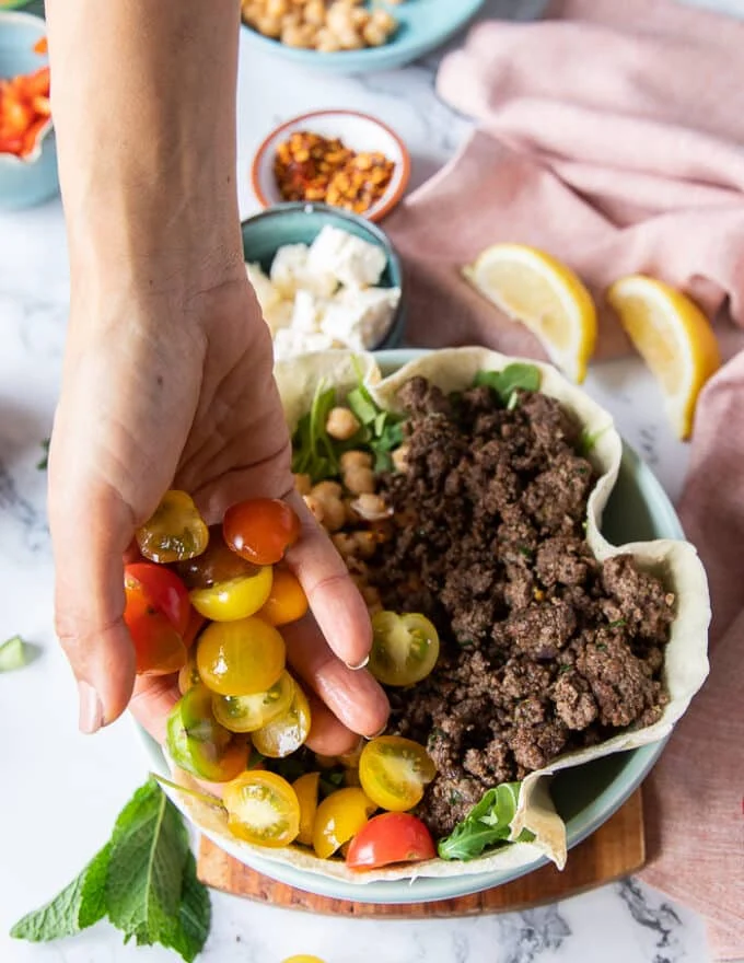 A hand adding in some cut tomatoes to layer the pita bowls as part of the many toppings