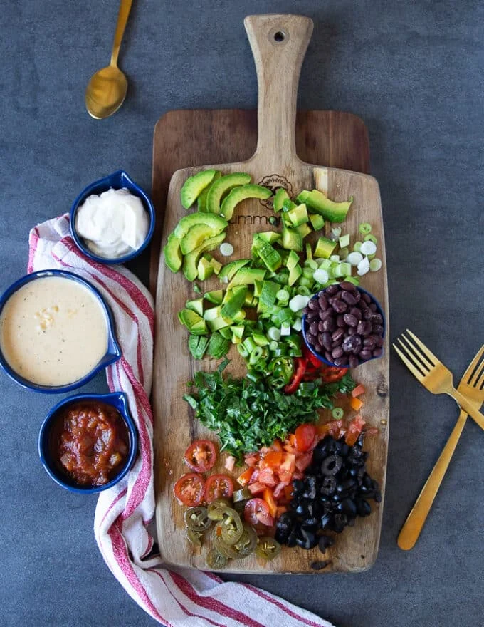 Many toppings in small bowls for nacho fries including taco beef, beans, nacho cheese sauce salsa, avocado, diced onions, tomatoes, cilantro, sour cream and pickled jalapeno