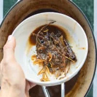 A hand holding a small plate of the extra whole tough spices and fresh sprigs after braising the lamb