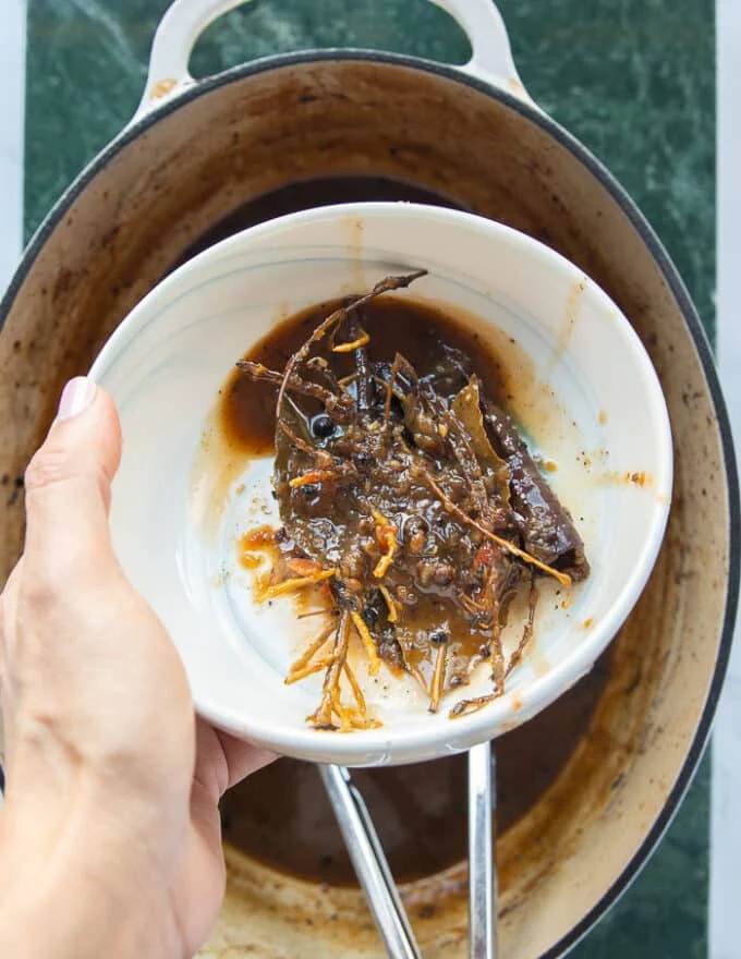 A hand holding a small plate of the extra whole tough spices and fresh sprigs after braising the lamb