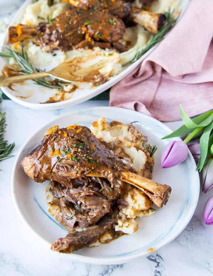 braised lamb shanks in a small plate with some mashed potatoes and broken up by a fork to show close up of how tender and soft the lamb is