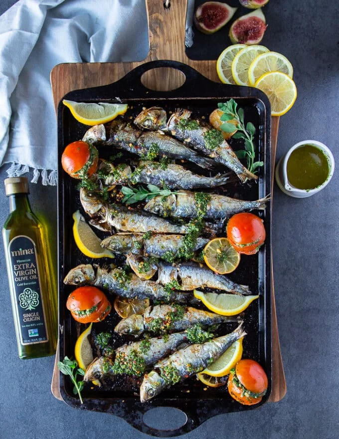 final baked sardines on a board with chermoula sauce, some lemon slices, chermoula stuffed tomatoes and olive oil