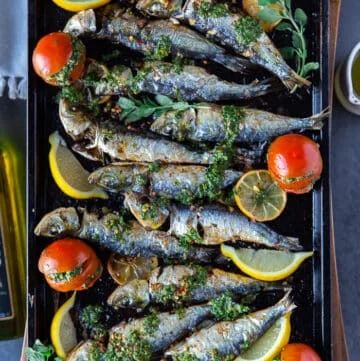 Baked sardines on a baking sheet with lemon slices, a drizzle of chermoula sauce and some stuffed tomatoes on the side