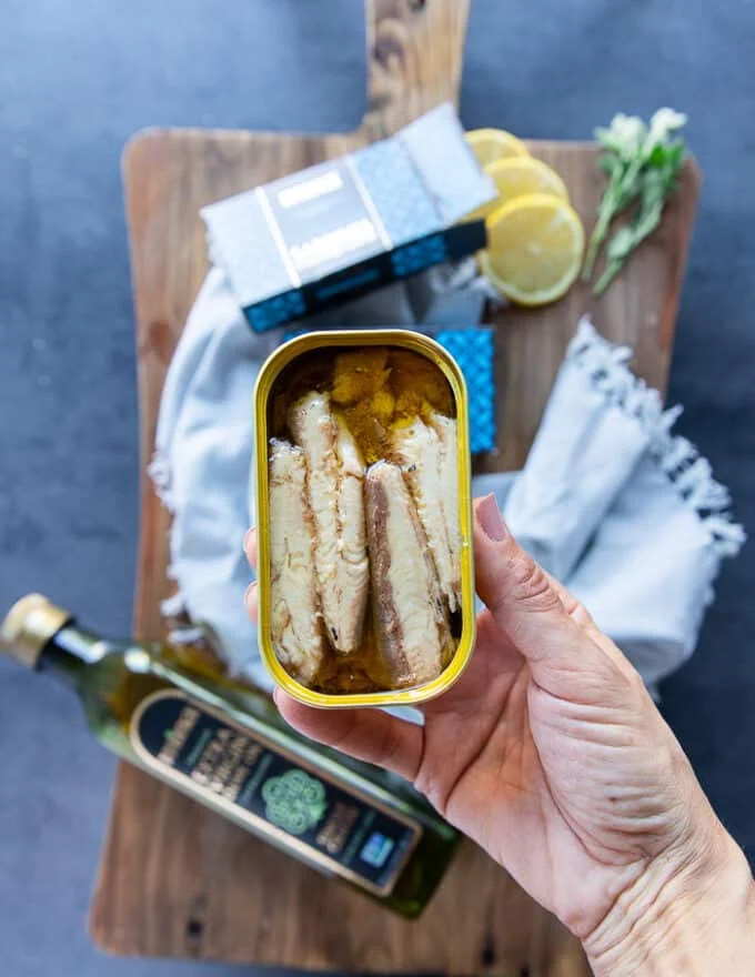 A hand showing a can of opened canned sardines showing the fillets of canned sardines inside the can with olive oil