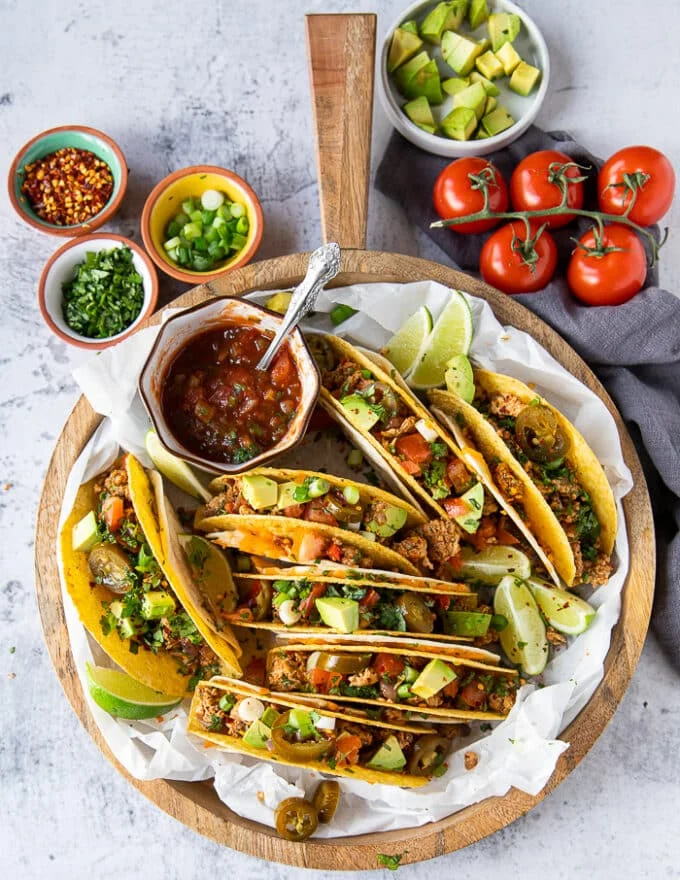 A board of ground chicken tacos loaded with toppings like avocado, green onions, salsa, cilantro and lime wedges