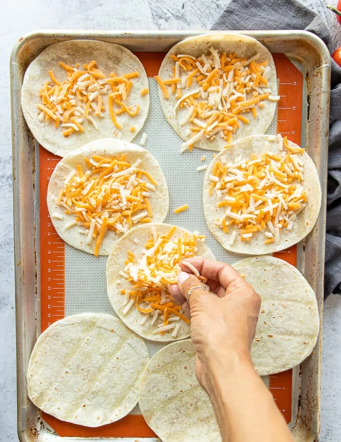 A hand sprinkling a generous amount of cheese on the soft shell tortillas on a baking sheet ready to melt in the oven
