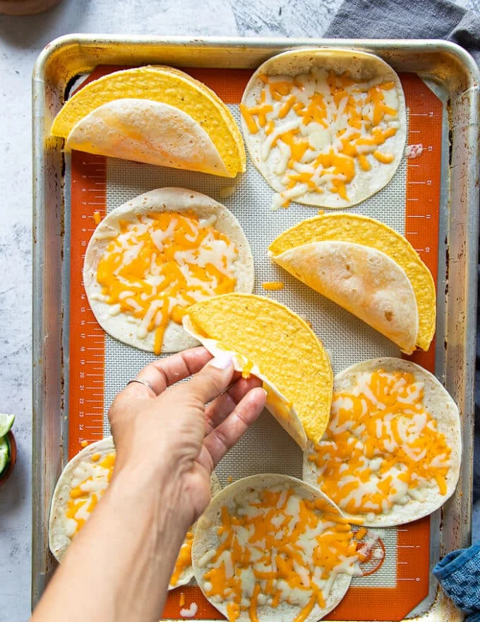 Once the cheese melted on the same baking sheet, a hand placing a hard shell tortilla and sticking it to the soft cheesy shell by pressing it down.