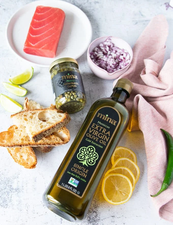 Ingredients to make tuna carpaccio including olive oil, lemon/lime juice, capers, onions, seasoning and green chilies