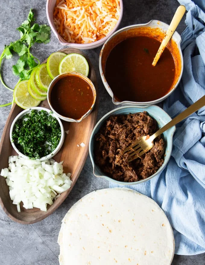 Ingredients to make birria quesadillas including: tortillas, birria meat, birria stew, cheese, cilantro, white onions chopped, jalapenos