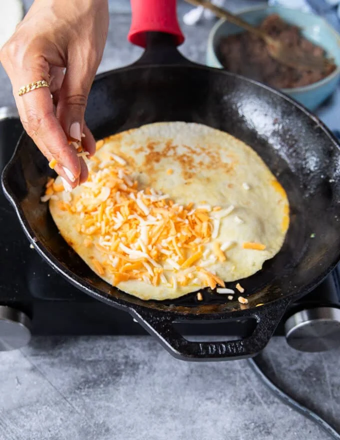 A hand scattering some shredded cheese al over the tortillas to cover it (while still int he pan)