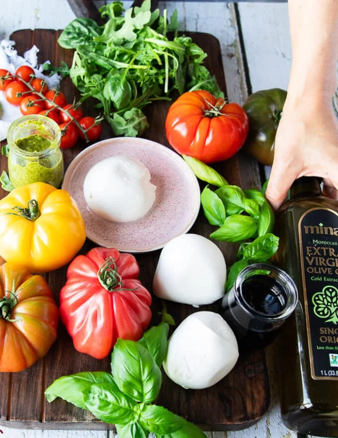 Caprese sandwich ingredients on a wooden board including fresh mozzarella , heirloom tomatoes, fresh basil leaves, burrata cheese, basil pesto, balsamic syrup and olive oil