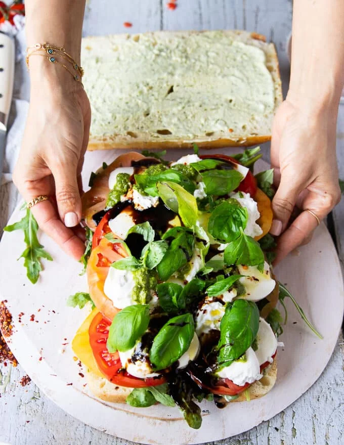 Basil leaves are added over the cheese in the caprese sandwich and a hand drizzling some balsamic syrup