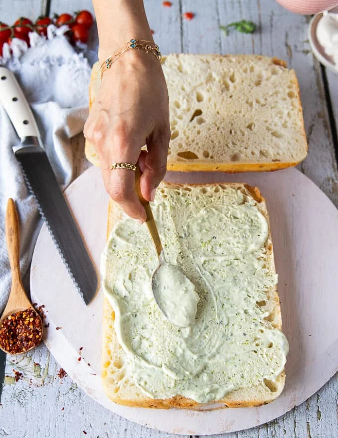 A hand spreading a layer of basil mayo over the bread to start making caprese sandwich