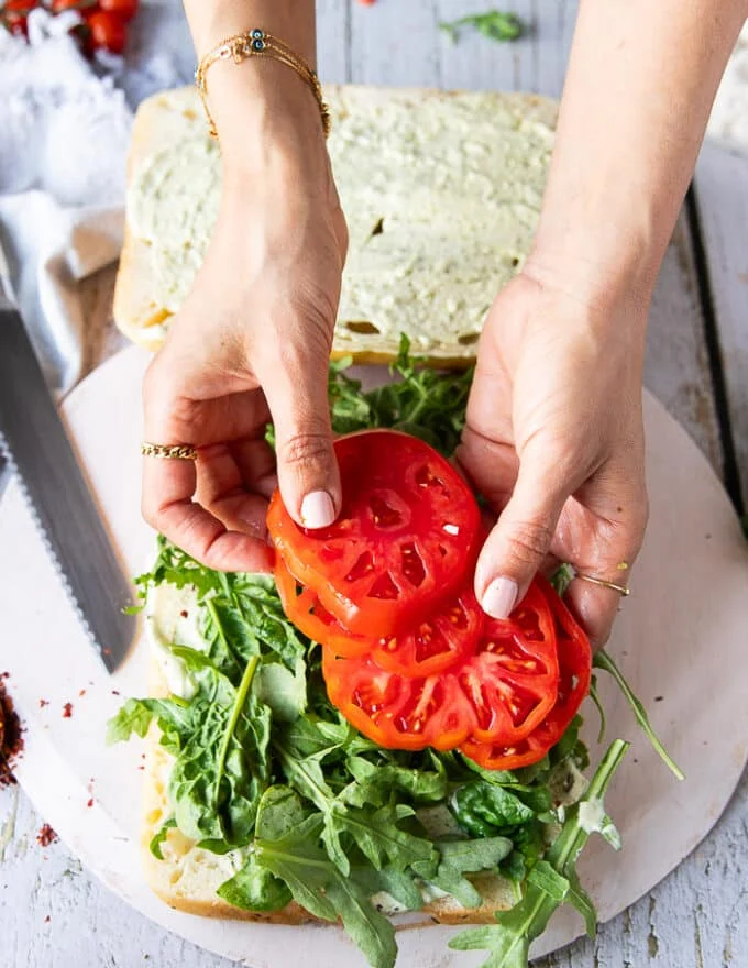 A hand assembling the sandwich starting with arugula, basil leaves and now adding the heirloom tomatoes