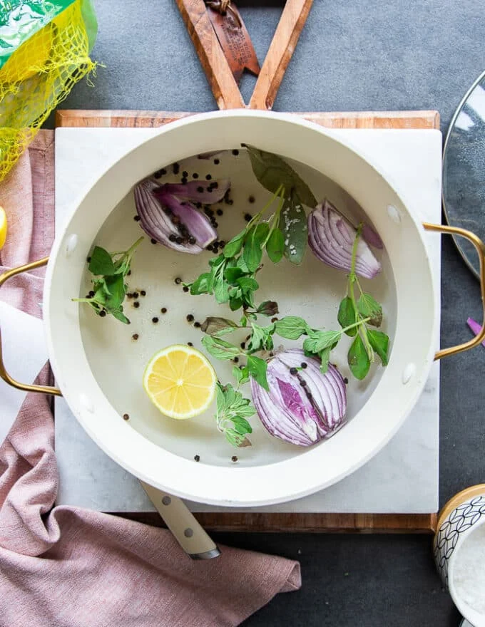 A pot filled with water, onion halves, lemon slices, fresh herbs, bay leaves, whole peppercorn and seasoning on the stove ready to boil for the octopus