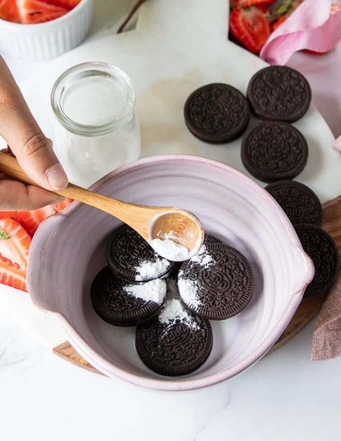 A hand pouring the baking powder over the oreo and milk in a a bowl