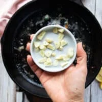 A hand holding sliced garlic ready to be added in to the same pan to make scmapi sauce