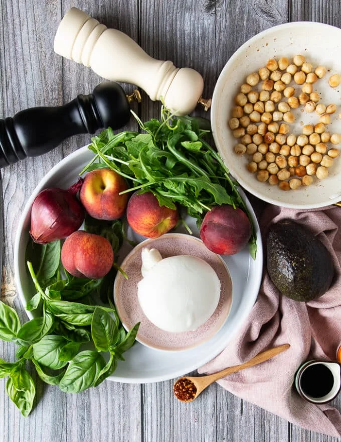 Ingredients for the peach burrata salad on a board including lots of fresh peaches, a burrata cheese, some basil leave, fresh arugula, onions, avocado and toasted hazelnuts