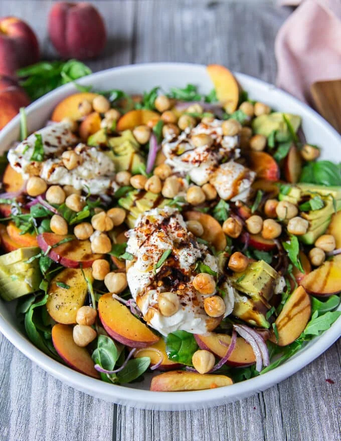 A close up of a plate of peach burrata salad showing the sliced peaches, the burrata cheese broken up, some basil leaves, avocados, sliced onions and a balsamic dressing