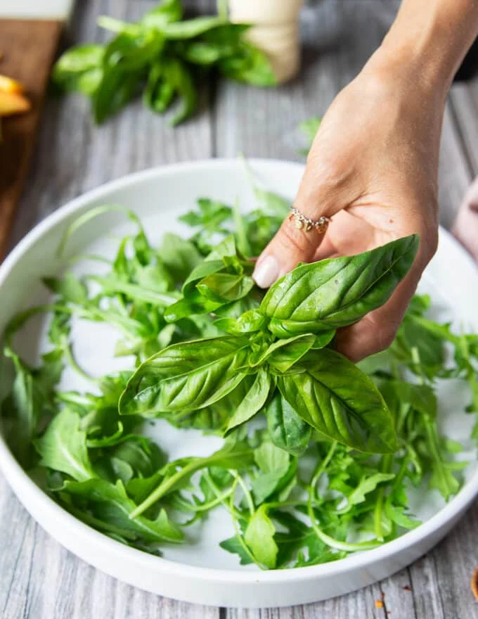 A hand layering the greens and fresh basil leaves at the bottom of the salad plate to make the salad recipe