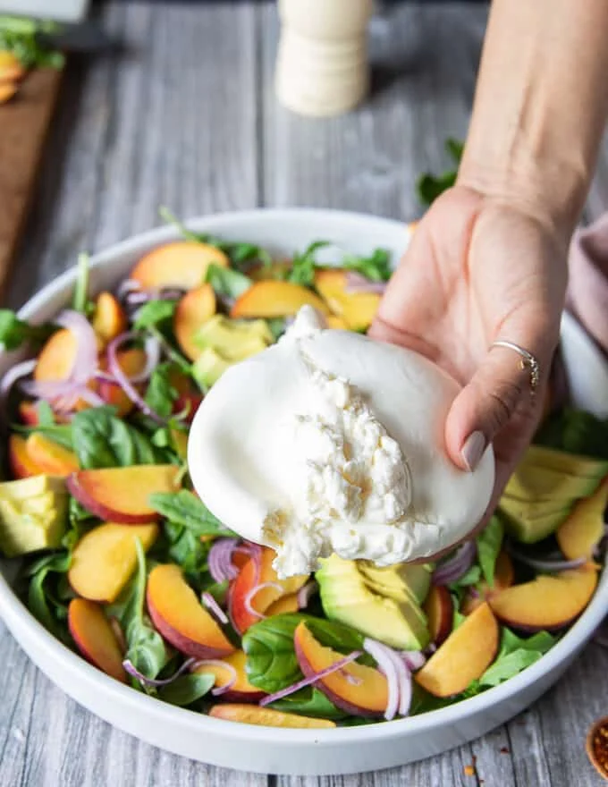 A hand holding the burrata cheese breaking it apart slightly over the peach salad