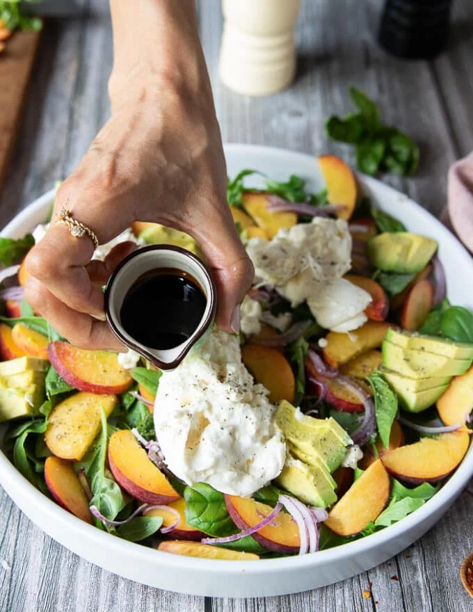 A hand pouring the balsamic syrup dressing over the assembled peach burrata salad