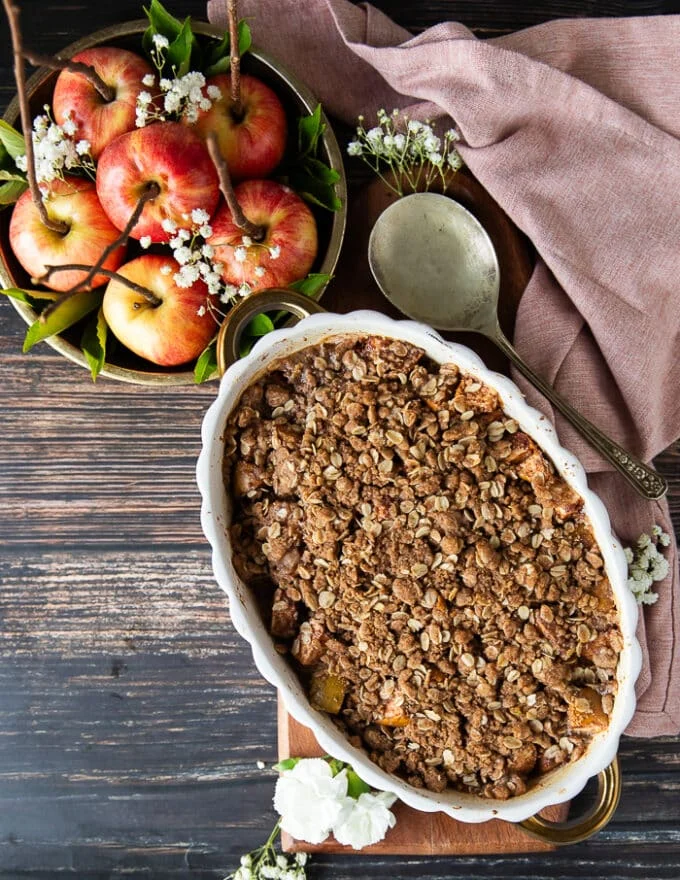 A baked apple crisp recipe in an oval dish and a large plate of apples on the side, along with a tea towel and a serving spoon