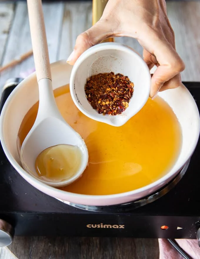 A hand adding in a small bowl of chilli flakes over the honey in a saucepan over medium heat
