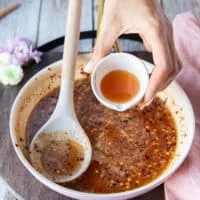 A hand pouring in a small bowl of apple cider vinegar to the sauce pan with honey and chili flakes off the heat