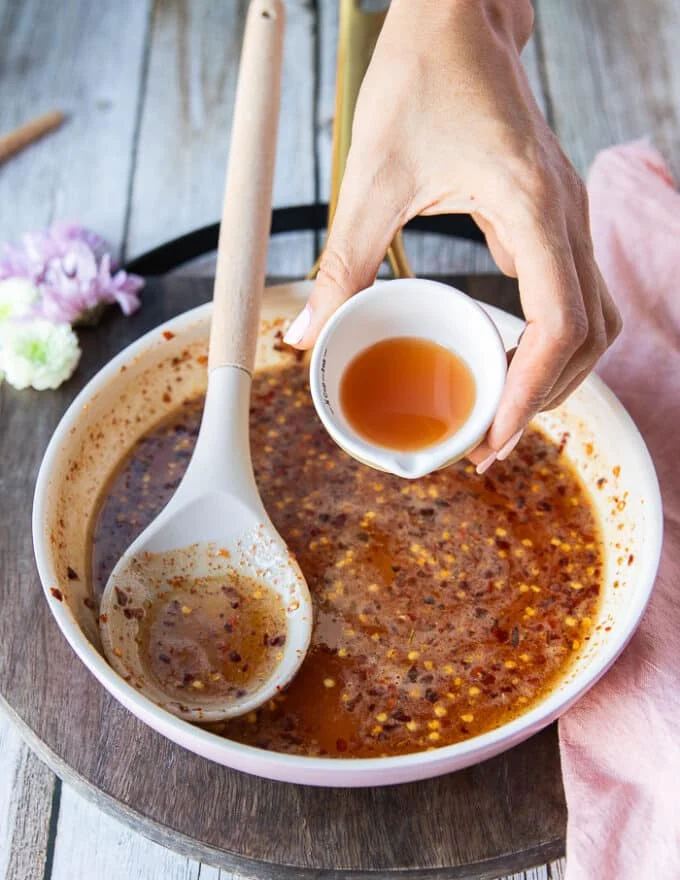 A hand pouring in a small bowl of apple cider vinegar to the sauce pan with honey and chili flakes off the heat