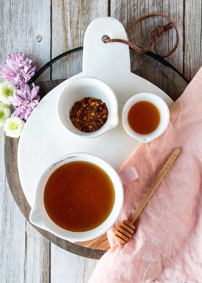 Ingredients for hot honey sauce including a bowl of honey, a bowl of chilli flakes, and a bowl of apple cider vinegar on a white marble