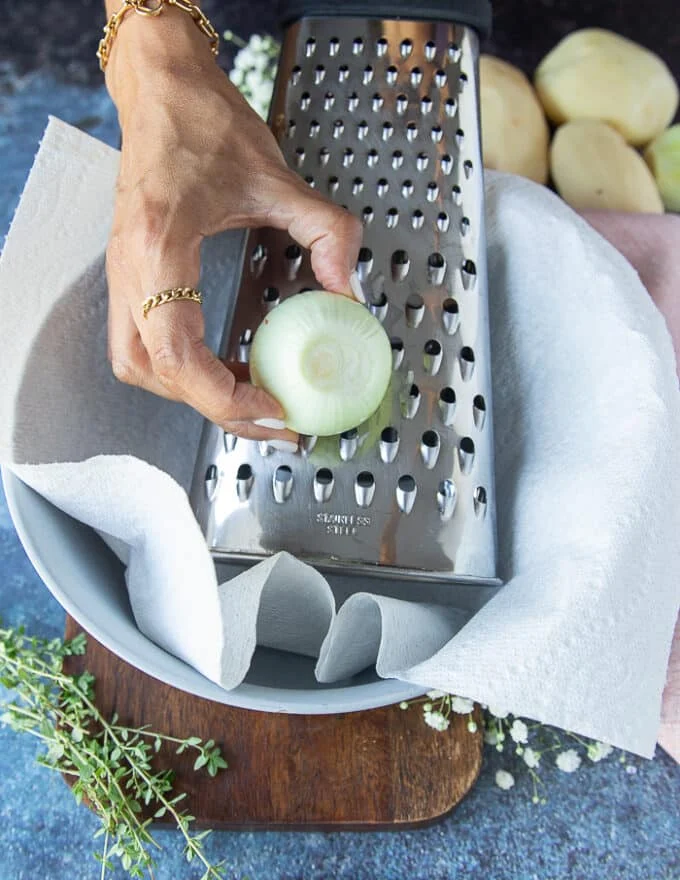 A hand holding onions and grating them using the same box grater over the bowl of grated potatoes lined with kitchen towel