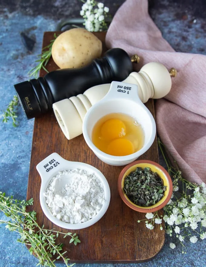A wooden board with ingredients for the best potato pancakes recipe including potatoes, eggs, onion, flour, salt and pepper and herbs