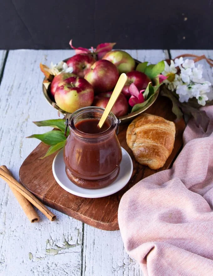 A kitchen table with a wooden board that has apple butter in a jar, some apples in the background, a tea towel, apple tree leaves, a pastry on the side on a plate set up for breakfast