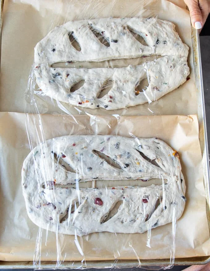The two fougasse breads on a baking sheet covered with plastic wrap and ready for the second rise before baking