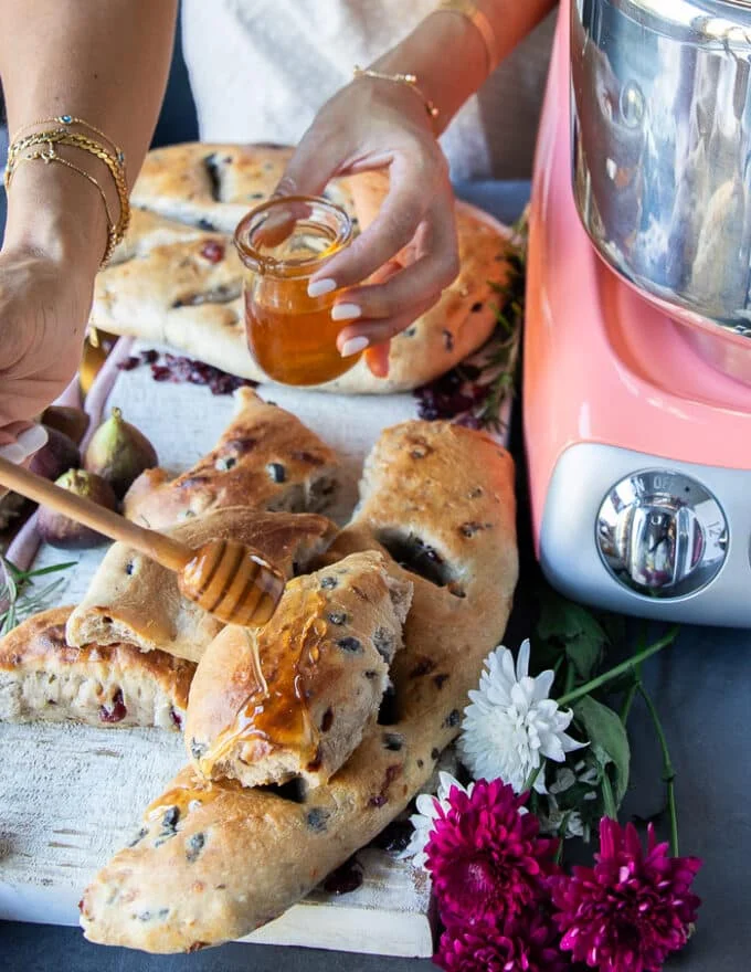 A hand drizzling honey on a loaf of fougasse bread over a cheese board