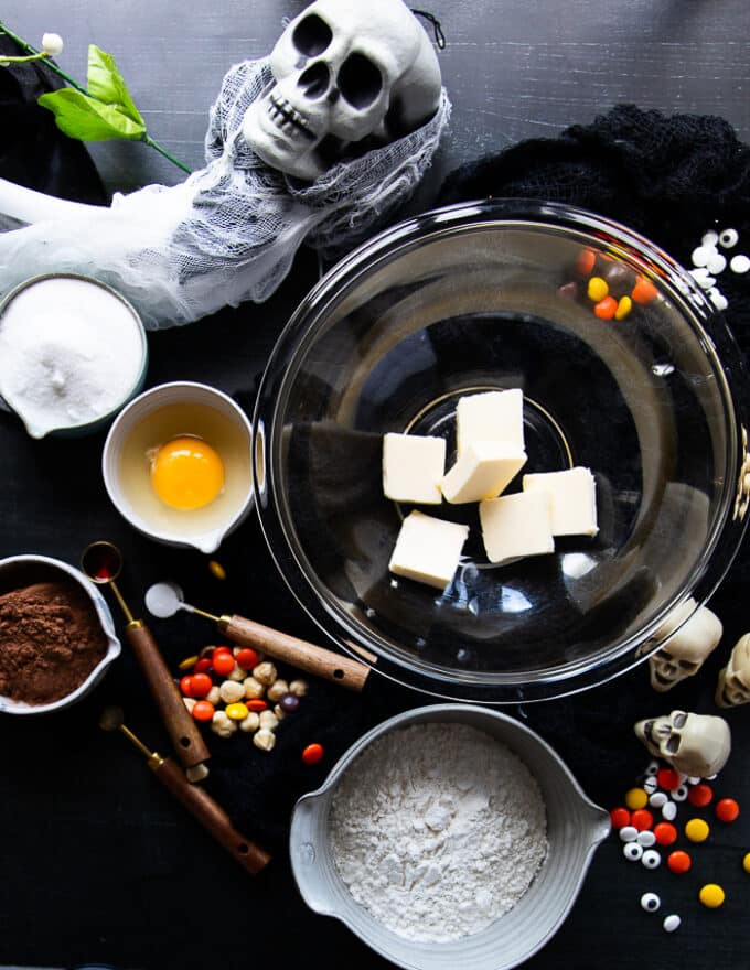 halloween cookies recipe ingredients including a bowl of soft butter, bowl of sugar, bowl with one egg, bowl with cocoa powder, baking soda in a spoon and a bowl of flour