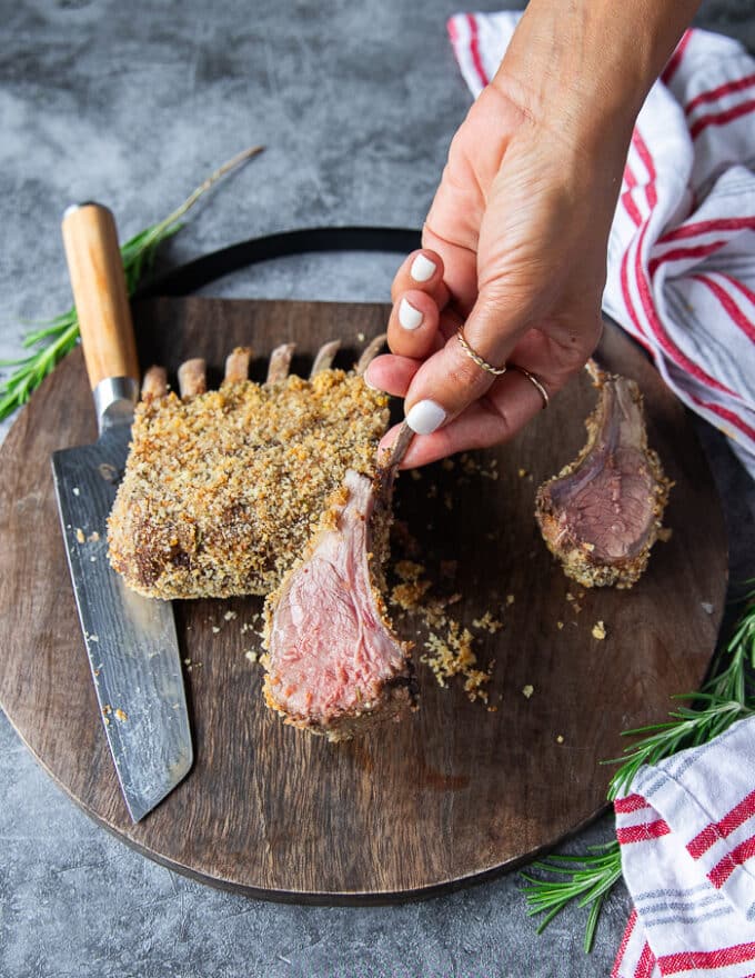A hand slicing a piece of the oven roasted lamb rack showing the medium rare meat and how tender it is