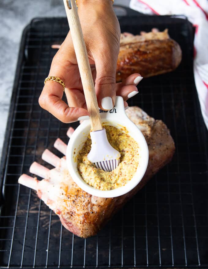 A hand holding a bowl of mustard and a brush to brush the surface of the lamb rack with mustard