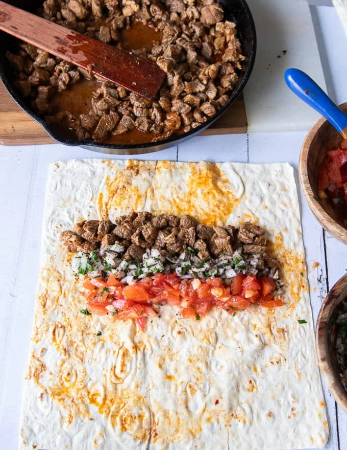 Thin Lavash bread on a wooden board with a spread of cooked meat, a thin spread of onion sumac salad and a thin spread of diced tomatoes. All in one layer, not mixed in